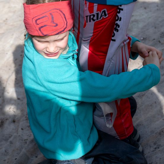 Ein junges Mädchen in einer türkisfarbenen Jacke mit einem roten Stirnband umarmt eine Person in roter und weißer Radsportausrüstung.