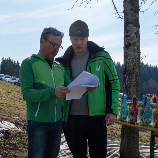 Zwei Männer stehen neben einem Baum, einer trägt eine grüne Jacke und der andere ein graues Hemd, beide schauen auf ein Stück Papier. In der Nähe steht ein Tisch mit verschiedenen Trophäenflaschen.