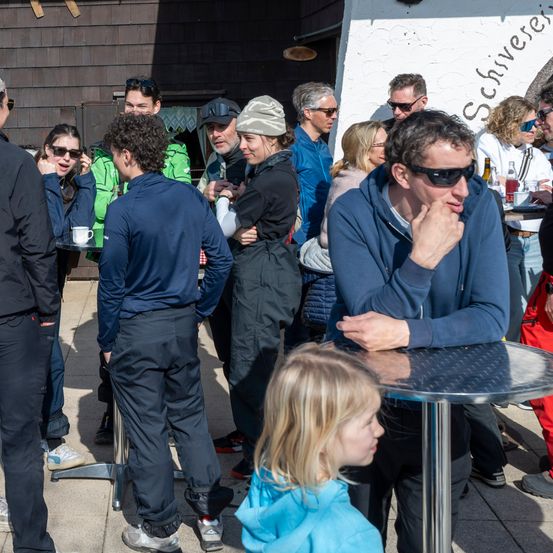 Eine Gruppe von Menschen versammelt sich draußen, ein Mann in einem blauen Mantel ruht sein Kinn auf seiner Hand. Ein Mädchen in einem blauen Mantel steht vor einem runden Tisch.