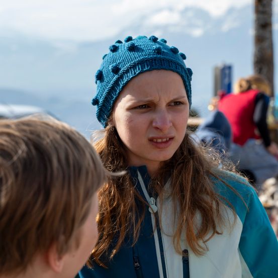 Eine Frau in einer blauen Mütze sieht besorgt aus, vielleicht im Gespräch mit einem Jungen mit braunen Haaren. Dahinter sind verschwommene Figuren zu sehen, und der Himmel ist teilweise bewölkt.
