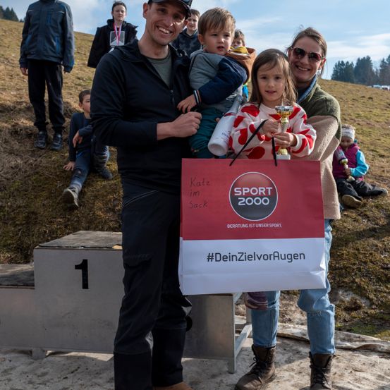 Eine Familie mit zwei Kindern hält einen Einkaufsbeutel mit dem Logo von Sport 2000. Sie stehen vor einem Podest, das mit der Nummer eins gekennzeichnet ist. Dahinter sitzen mehrere Kinder auf dem Boden, und eines hält einen Pokal.