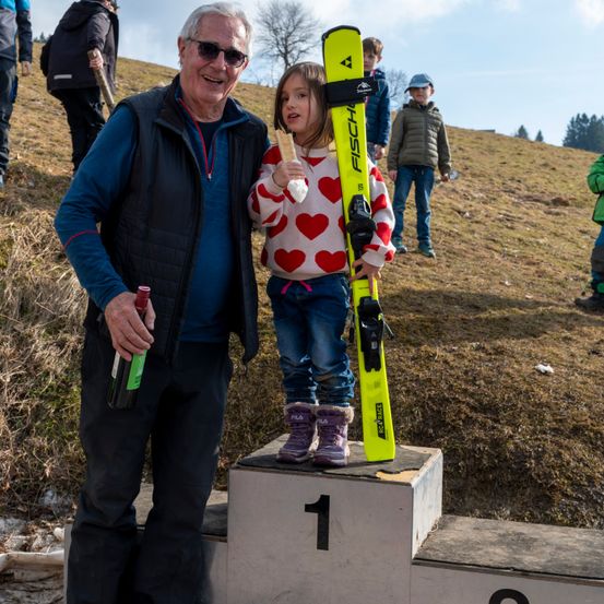 Ein älterer Mann mit Sonnenbrille steht auf einem Podest mit einem jungen Mädchen, das Skier hält. Sie lächeln beide. Hinter ihnen laufen mehrere Personen auf einem Grashügel.