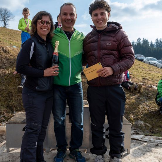 Drei Personen stehen lächelnd auf einem Podest. Der Mann in Grün hält eine Flasche und der Mann in Braun ein gelbes Papier. Hinter ihnen steht ein Junge mit einer Medaille auf einem Grashügel.