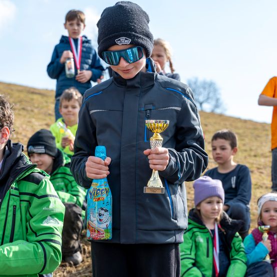 Ein junger Junge in einer schwarzen Jacke hält einen Pokal und eine blaue Flasche in einer Außenumgebung. Hinter ihm sitzen mehrere Kinder auf dem Boden, einige tragen Medaillen.
