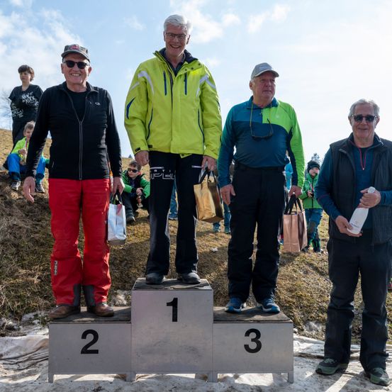 Drei Männer stehen auf einem Podium mit den Zahlen eins, zwei und drei. Sie lächeln und tragen Winterkleidung. Hinter ihnen stehen Menschen und schauen zu. Der Hintergrund ist ein Hügel mit Gras.