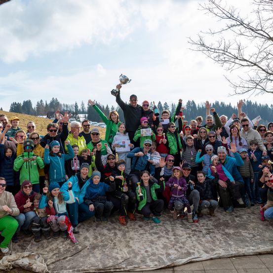 Eine große Gruppe von Menschen posiert für ein Foto, hält Pokale und trägt Winterkleidung. Ein Mann hält einen Pokal hoch, während andere jubeln. Der Hintergrund besteht aus Bäumen und einem bewölkten Himmel.