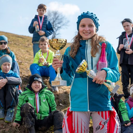 Ein junges Mädchen hält einen goldenen Pokal in der Hand, trägt einen blauen Hut und eine blaue Jacke und wird von Kindern auf einem Grasfeld umgeben, einige sitzen und andere stehen, unter einem blauen Himmel mit Wolken.