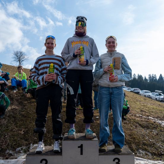 Drei junge Menschen stehen auf einem Podest mit Nummernschildern. Sie tragen Winterkleidung und halten Getränke in den Händen. Der erste Platz ist links, der zweite Platz in der Mitte und der dritte Platz rechts.