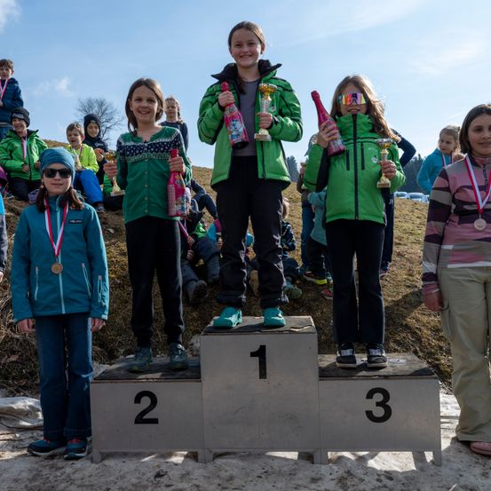 Eine Gruppe junger Mädchen steht auf einem Podest mit Medaillen und Pokalen. Eines Mädchen steht in der Mitte auf dem ersten Platz, und zwei andere stehen an den Seiten. Der Hintergrund zeigt eine Menge von Kindern, die auf dem Boden sitzen.