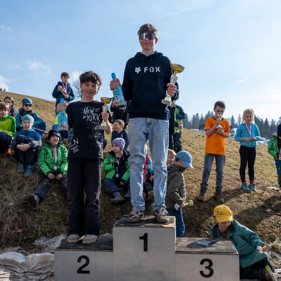 Zwei junge Jungen stehen auf einem Podest und halten Pokale. Einer trägt eine schwarze Fox-Kapuzenjacke, der andere ein New York T-Shirt. Sie sind von anderen Kindern umgeben, die Medaillen tragen und auf dem Gras sitzen. Bäume und ein klarer Himmel sind im Hintergrund.