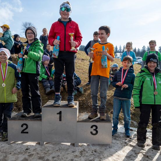 Mehrere Kinder stehen auf einem Podium mit den Nummern 1, 2 und 3, halten Trophäen und Medaillen. Sie tragen Winterjacken und Turnschuhe. Dahinter sehen weitere Kinder und Erwachsene zu.