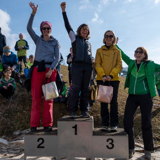 Vier Frauen stehen auf einem Podest und feiern. Eine hält eine Trophäe. Zuschauer sitzen hinter ihnen auf dem Gras. Der Himmel ist teilweise bewölkt.