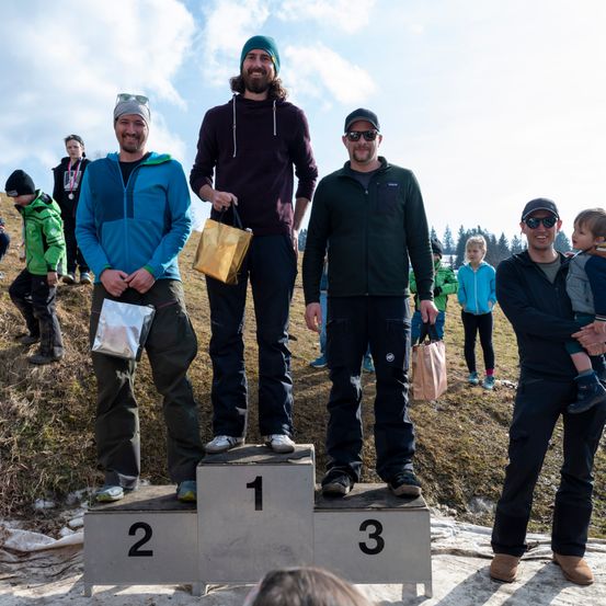 Drei Männer stehen auf einem Podest mit den Nummern 1, 2 und 3. Sie tragen Winterkleidung und halten Taschen. Hinter ihnen stehen einige Leute, darunter ein Kind in den Armen eines Mannes. Sie stehen auf einem Grashügel unter einem bewölkten Himmel.
