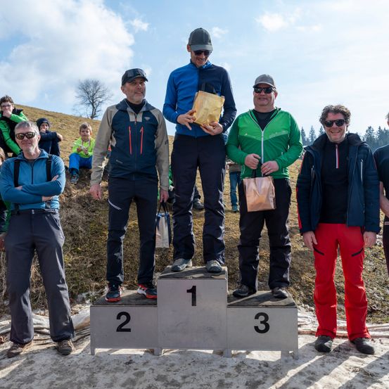 Die Gewinner eines Rennens stehen auf einem Podium mit den Nummern 1, 2 und 3. Der Mann in der Mitte hält einen Pokal. Sie tragen alle eine Sonnenbrille.