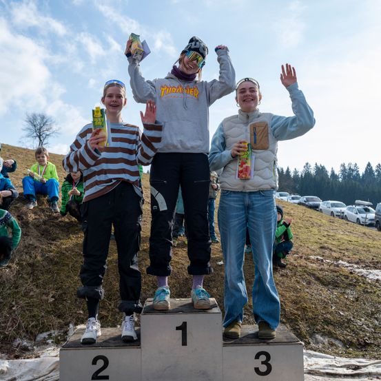 Drei junge Personen stehen auf einem Podest, jede hält eine Saftpackung. Die Person in der Mitte trägt eine graue Kapuzenjacke und schwarze Hose. Die anderen tragen ein gestreiftes Shirt und blaue Jeans. Hinter ihnen sitzen eine Gruppe von Menschen auf dem Rasen.