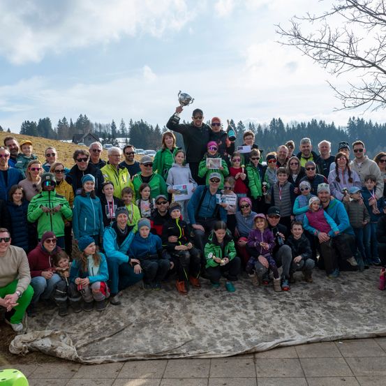 Eine große Gruppe von Menschen, darunter Erwachsene und Kinder, versammelt sich für ein Foto. Sie tragen Winterkleidung und einige halten Pokale. Ein Mann in der Mitte hält einen Pokal über seinem Kopf. Im Hintergrund befindet sich ein Hügel mit Bäumen.