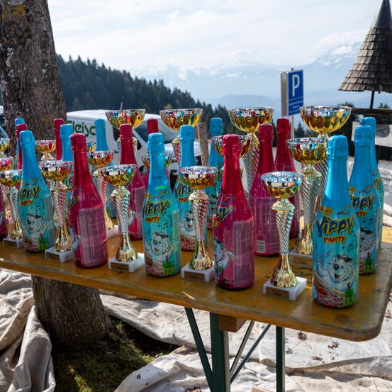 Ein Tisch mit verschiedenen Trophäen, darunter einige in Funkelflaschendesigns eingewickelt, ist im Freien aufgestellt. Im Hintergrund befinden sich Berge und ein klarer Himmel.