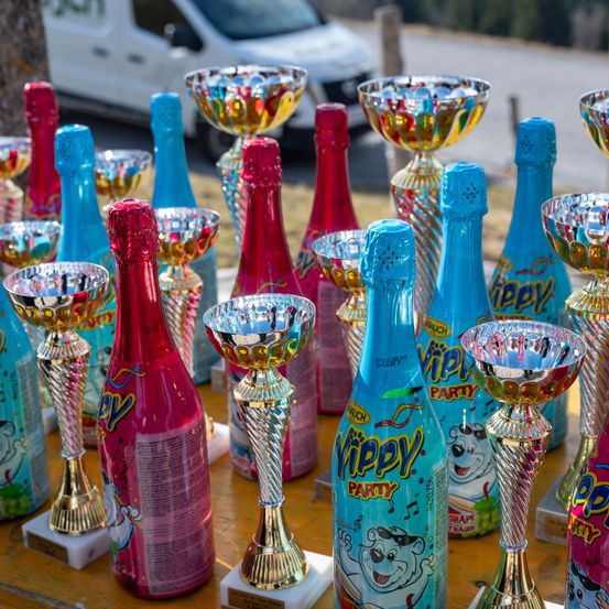 Mehrere farbenfrohe Pokale und Yippy Party Flaschen sind auf einem Tisch ausgestellt. Ein weißer Lieferwagen ist im Hintergrund geparkt.