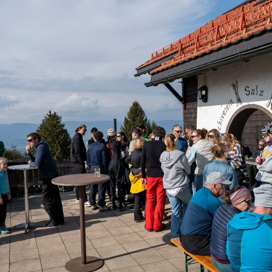 Viele Menschen haben sich um eine kleine Bar in einem Außenbereich versammelt, möglicherweise in einem Skigebiet. Sie stehen und sitzen in einer Reihe. Das Gebiet ist von Bäumen umgeben.