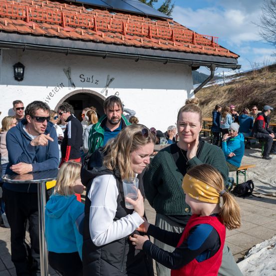 Eine Gruppe von Menschen steht vor einem Gebäude mit einem roten Dach. Solarpanele sind auf dem Dach installiert. Zwei Frauen unterhalten sich mit einem Mädchen vor ihnen. Einige Leute sitzen auf Bänken, andere stehen herum.