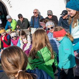 Eine Gruppe von Menschen, einschließlich Kindern, ist um einen Tisch herum im Freien versammelt. Sie tragen Winterkleidung und einige haben Sonnenbrillen auf. Flaschen stehen auf dem Tisch.