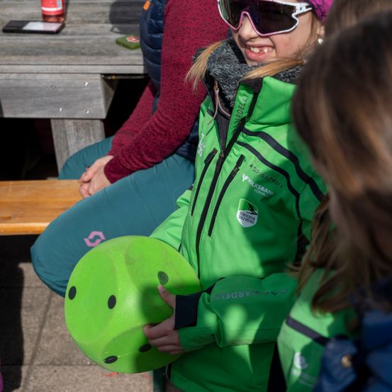 Ein Mädchen in einer grünen Jacke hält ein grünes Spielzeug und lächelt. Hinter ihr sitzt eine Person auf einer Bank in einem burgunderroten Pullover. Vor ihnen steht ein Holztisch mit einer Flasche und einem Handy.