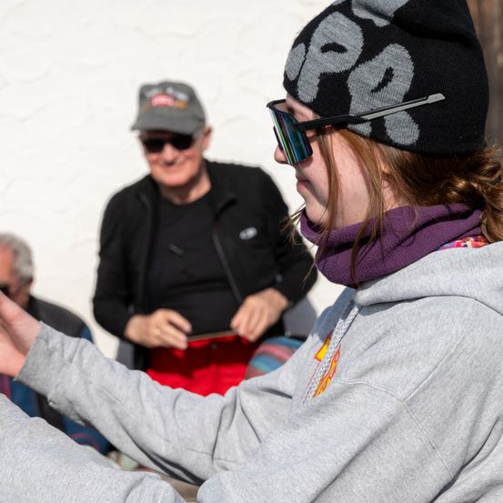 Eine Frau mit grauer Kapuzenjacke und Sonnenbrille schaut weg, mit einem Mann in einer Kappe hinter ihr. Ein anderer Mann ist im Hintergrund teilweise sichtbar.