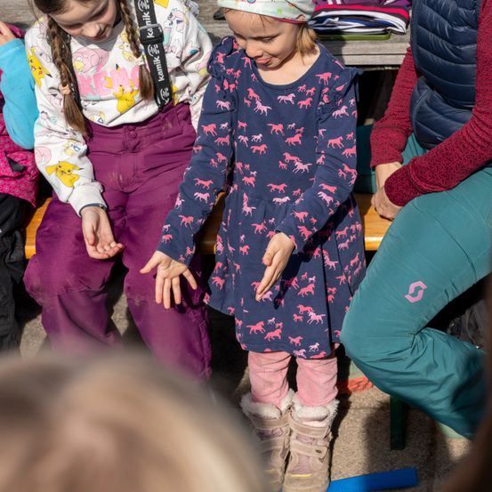 Ein kleines Mädchen mit einem Pferde-Thema-Kleid steht zwischen zwei anderen Kindern, eines trägt ein Pokemon-Shirt. Sie sitzen auf einer Bank.