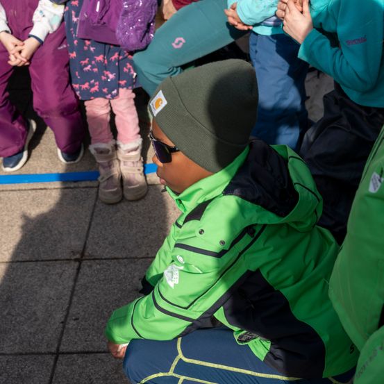 Ein junger Junge in einer grünen Jacke und Mütze sitzt auf dem Boden, umgeben von anderen Kindern in Winterkleidung.