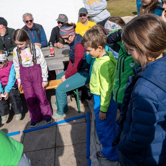 Eine Gruppe von Erwachsenen und Kindern sitzt draußen um einen Holztisch. Ein Mädchen in lila Hosen steht in der Nähe des Tisches und ein Junge in grüner Hose steht vor ihr. Eine Frau mit Stirnband sitzt neben ihnen.