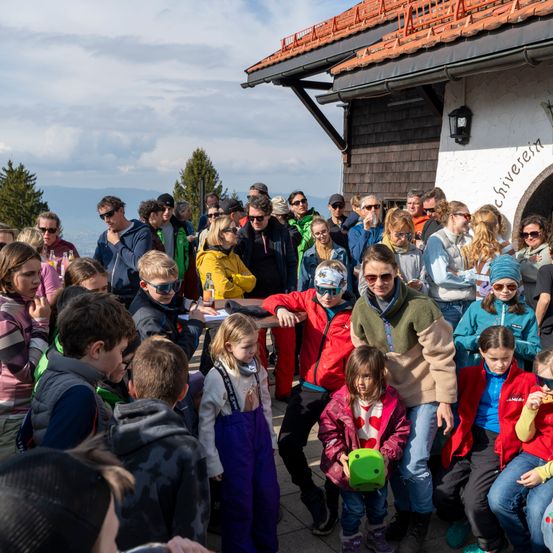 Eine Gruppe von Menschen versammelt sich vor einem Chalet. Erwachsene und Kinder in Winterkleidung, einige mit Brille, stehen um einen Tisch mit Essen. Ein Kind hält einen grünen Würfel.