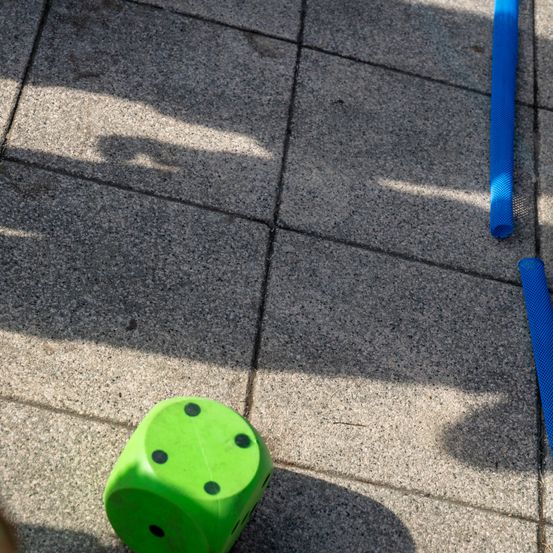 Ein grüner Würfel mit schwarzen Punkten liegt auf einem gefliesten Boden mit Schatten und blauen Röhren in der Nähe.