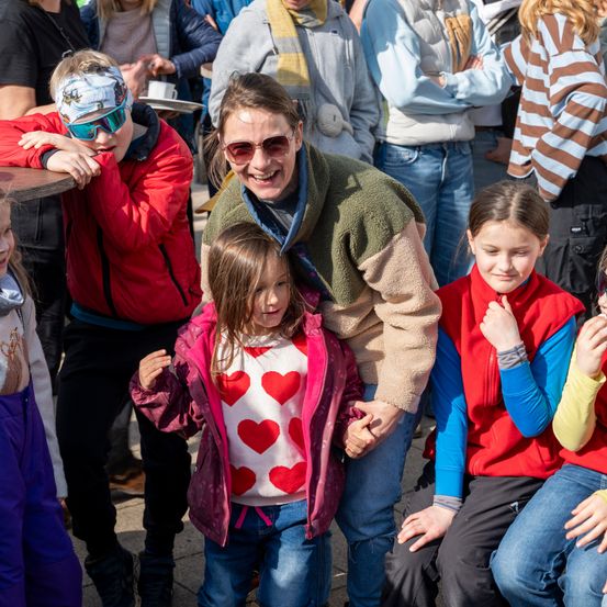 Eine Gruppe von Kindern und Erwachsenen ist draußen, wobei eine Frau die Hand eines Kindes hält. Das Kind trägt ein herzförmiges Shirt. Die Frau lächelt und andere im Hintergrund beobachten.