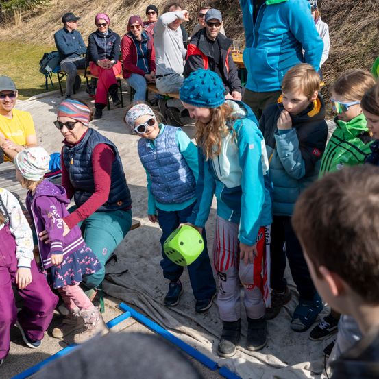 Eine Gruppe von Menschen, einschließlich Kindern, versammelt sich im Freien, einige sitzen auf Bänken, andere stehen. Ein Kind hält einen grünen Würfel, und mehrere schauen zu.