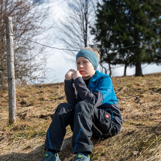 Ein junger Junge in einer blauen Jacke und einem Stirnband sitzt auf einem Grashügel und schaut nachdenklich mit den Händen am Kinn. Hinter ihm ist ein Zaun und Bäume im Hintergrund.