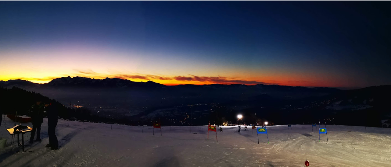 Eine Skipiste mit Flaggen und beleuchteten Stangen unter einem Dämmerungshimmel mit Bergen im Hintergrund.