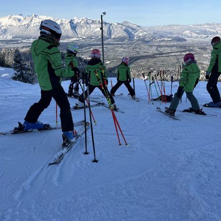Eine Gruppe von Skifahrern, die grüne Jacken und Helme tragen, rutschen einen verschneiten Berg hinunter. Sie halten Skistöcke und stehen in einer Reihe.