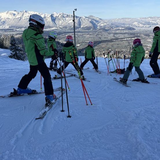 Eine Gruppe von Skifahrern, die grüne Jacken und Helme tragen, rutschen einen verschneiten Berg hinunter. Sie halten Skistöcke und stehen in einer Reihe.