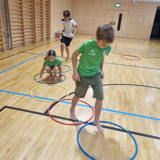 Drei Kinder spielen mit Hula-Hoops auf einem Indoor-Spielfeld. Ein Mädchen kauert in der Mitte, und zwei Jungen stehen.