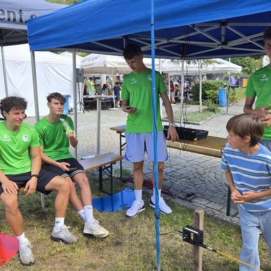 Vier junge Männer in grünen T-Shirts sitzen unter einem blauen Zelt. Einer von ihnen steht und hält ein Handy. Ein Junge in einem gestreiften T-Shirt steht in der Nähe und schaut sie an.