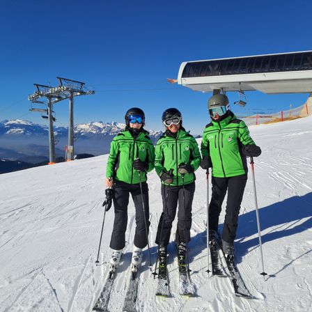 Drei Skifahrer in grünen Jacken stehen auf einem verschneiten Berg, lächeln und halten Skistöcke. Hinter ihnen ist eine Skiliftstation mit einer Gondel zu sehen, und die schneebedeckten Berge bilden einen malerischen Hintergrund.