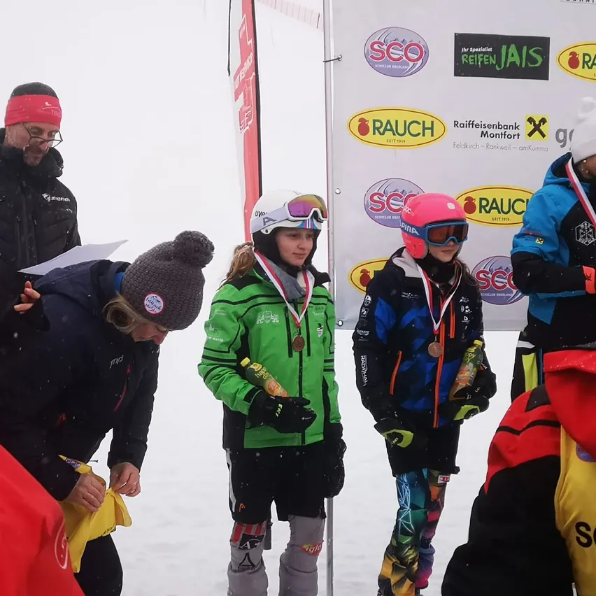 Drei junge Skifahrer in bunter Skiausrüstung mit Medaillen stehen auf einem verschneiten Feld. Sie sind von Erwachsenen umgeben, einer hält ein Stück Papier. Ein Banner mit Logos und Text steht hinter ihnen.