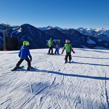Vier Kinder sind auf einem verschneiten Berg beim Skifahren. Ein Kind trägt eine grüne Jacke, die anderen sind in Blau. Der Schnee ist gut präpariert und der Berg ist im Hintergrund.