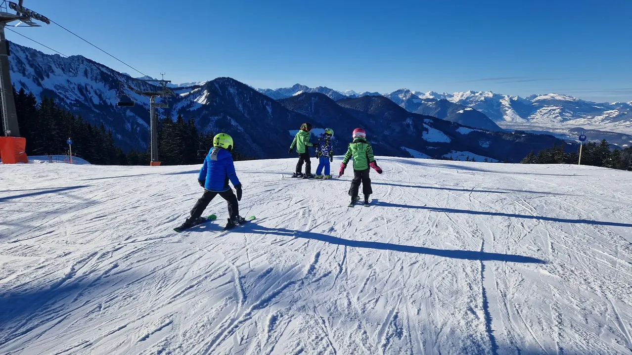 Vier Kinder sind auf einem verschneiten Berg beim Skifahren. Ein Kind trägt eine grüne Jacke, die anderen sind in Blau. Der Schnee ist gut präpariert und der Berg ist im Hintergrund.