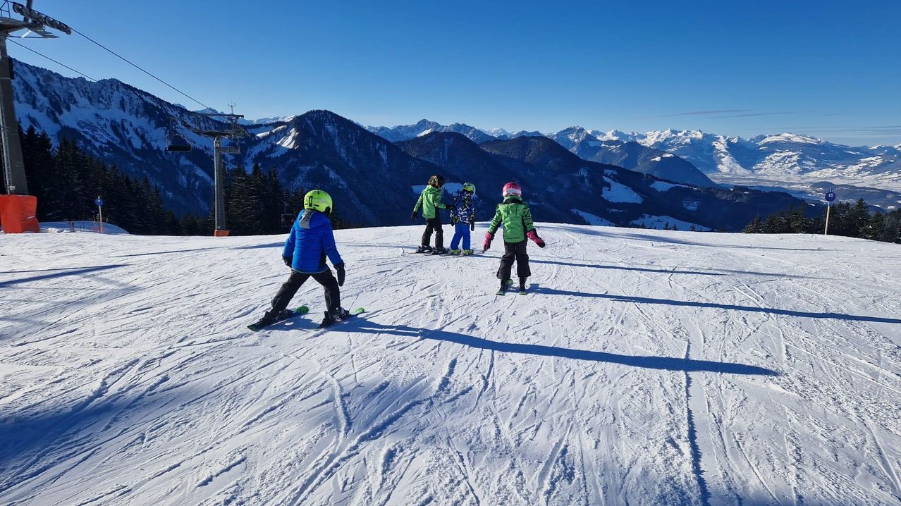 Vier Kinder sind auf einem verschneiten Berg beim Skifahren. Ein Kind trägt eine grüne Jacke, die anderen sind in Blau. Der Schnee ist gut präpariert und der Berg ist im Hintergrund.