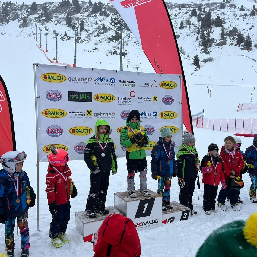 Eine Gruppe von Kindern steht auf einem verschneiten Podest und hält Medaillen. Sie tragen Skiausrüstung und Helme. Dahinter zeigt ein großes Banner Logos und Text. Im Hintergrund sind schneebedeckte Berge und Skilifte zu sehen.