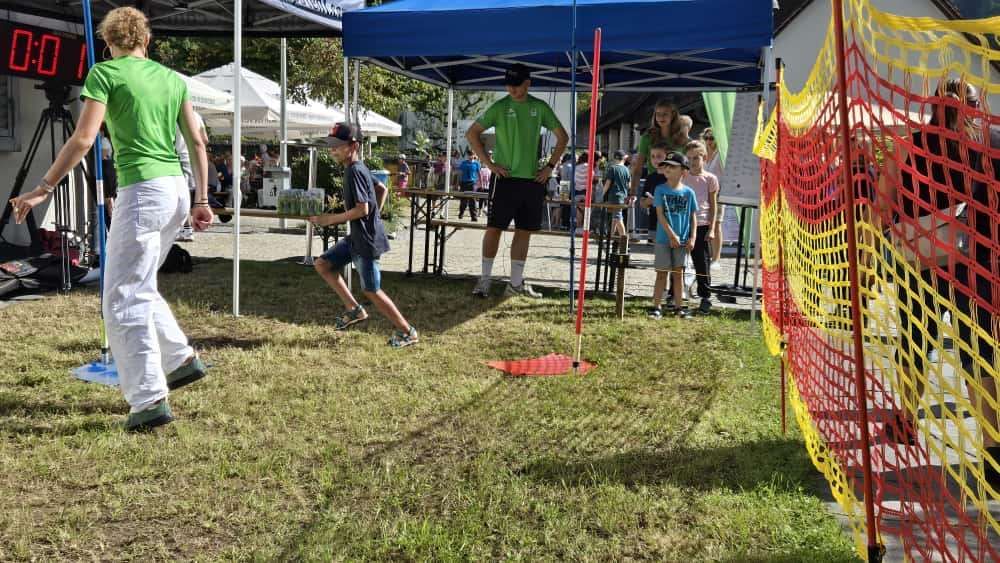 Ein Junge läuft auf einem Rasenfeld und zielt auf ein Ziel, während ein Erwachsener zusieht. Andere Kinder und Erwachsene stehen in der Nähe eines blauen Zeltes.