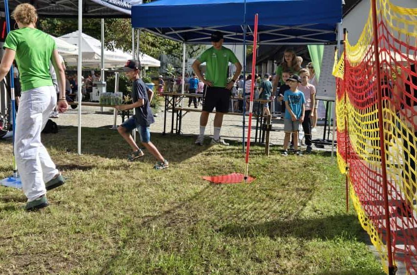 Ein Junge läuft auf einem Rasenfeld und zielt auf ein Ziel, während ein Erwachsener zusieht. Andere Kinder und Erwachsene stehen in der Nähe eines blauen Zeltes.