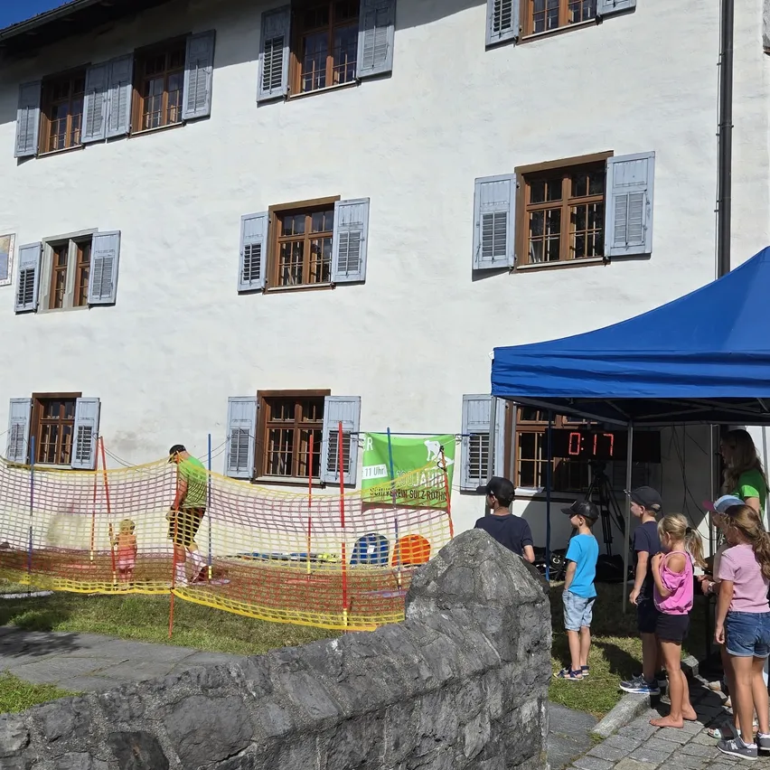 Eine Gruppe von Kindern beobachtet einen Mann in einem grünen Shirt und Hut, der einen Ball wirft. Ein blaues Zelt mit einem digitalen Timer steht vor einem Gebäude mit weißen Wänden und braunen Fensterläden.