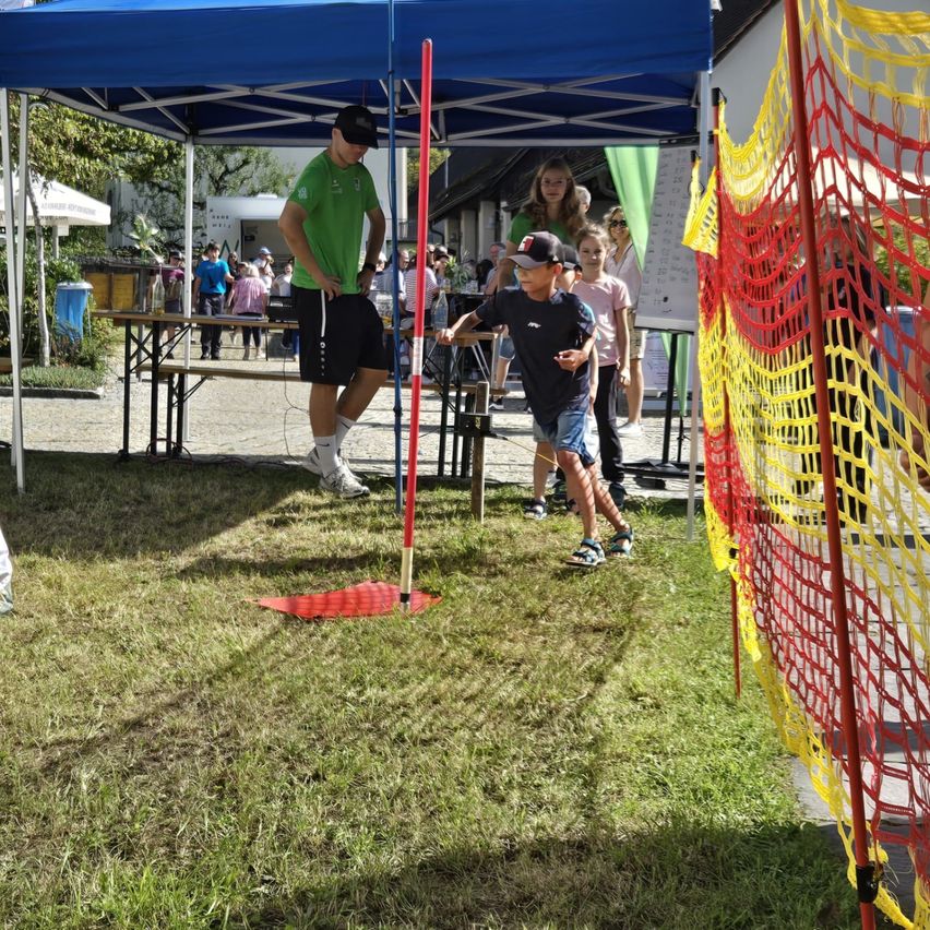 Ein Junge wirft einen Speer auf einem Rasenfeld unter einem blauen Zelt, wobei Zuschauer zusehen, einschließlich eines Mannes in einem grünen T-Shirt.